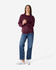 A woman stands wearing a burgundy Gildan Heavy Blend adult crewneck sweatshirt and blue jeans. The sweatshirt features a logo.