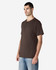 A man stands wearing a brown Gildan Heavy Cotton Adult T-Shirt. The shirt has a logo on it and a relaxed fit.