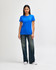 A woman stands wearing a blue Gildan Softstyle ladies' t-shirt and denim pants, paired with white sneakers.