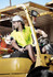 A man wearing a high-visibility yellow short sleeve polo sits on a forklift, wearing a white safety helmet.