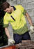 A bright yellow short sleeve polo shirt with a geometric pattern, worn by a man in work gloves, set against a stone wall.