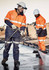 Two workers in high-visibility orange jackets and helmets are cleaning a surface, wearing gloves and using tools.