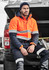 A man wearing an orange and navy unisex hi-vis softshell jacket sits on the back of a vehicle.