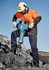 A man in an orange and black jacket and ear protection operates a blue jackhammer on rocky terrain.