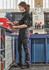 A woman in black rugged cargo pants stands at a workshop bench, engaging with tools and equipment. The pants feature pockets and a logo.