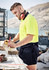 A man in a high-visibility yellow shirt and navy rugby shorts measures a wooden plank at a construction site.