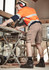 A man wearing beige cargo shorts and a high-visibility orange shirt works with power tools in a construction setting.
