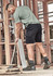 A man in a grey t-shirt and black shorts works on a construction site, holding a broom and wearing sturdy boots.