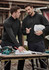 Two men in black unisex 1/4 zip brushed fleece pullovers discuss plans at a worktable outdoors, with tools visible.