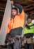 Unisex hi-vis full zip hoodie in orange and black, featuring a logo, worn by a model on a construction site.