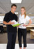 A man in a black polo shirt and a woman in a white polo shirt hold a folder in an office setting.