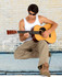 A man wearing a white cotton rib singlet and sunglasses plays an acoustic guitar, sitting against a brick wall.