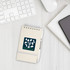 A beige notepad with a spiral binding and a pen, featuring a logo on the front, on a white desk.