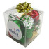 A clear cube filled with assorted Christmas chocolates wrapped in red, green, and silver foil, topped with a gold bow.
