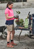 A woman in a pink and navy polo shirt wears beige cargo shorts, holding plants beside gardening tools and a wheelbarrow.