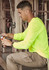 A man works in a construction setting, wearing a neon yellow long sleeve tee and beige cargo pants, using pliers.