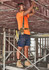 A worker in an orange shirt and navy cargo shorts uses a power tool on a construction site.