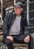 A man wearing a black unisex packable rain jacket sits on scaffolding. The jacket features a hood and has a logo.