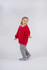 A young girl in a red v-neck fleece jumper and grey pants, smiling and standing against a light background.