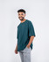 A man in a teal unisex oversize easy-fit tee stands smiling, paired with grey trousers. The shirt has a logo.