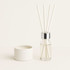 A vanilla reed diffuser with white reeds, housed in a glass bottle with a silver collar, alongside a white container.