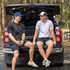 Two individuals wearing caps sit on the tailgate of a black vehicle, surrounded by trees. One cap is blue, the other is white.