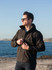 A man wearing a black soft shell jacket stands by the water, adjusting his jacket with sunglasses on.