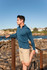 A man in a blue long-sleeve top and beige shorts stretches by the water, with rocky terrain in the background.
