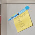 A blue plastic pen shaped like a house is attached to a fridge, alongside a yellow shopping list note.