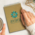 A kraft notepad with a spiral binding and a hand holding a pen, beside a cup of coffee. It features a logo on the cover.