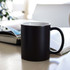 A ceramic mug in matte black with a white interior, placed on a table next to magazines. It features a handle and a logo.