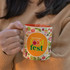 A coffee mug with a vibrant orange interior and a multicoloured fruit-themed exterior, featuring a logo.