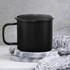 A black enamel metal cup with a handle, placed on a marble coaster and surrounded by a grey knitted item.