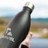 A drink bottle in matte black with a silver lid and a logo, held against a blurred outdoor background.
