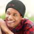 A man smiles while wearing a dark green cable knit beanie with a logo, set against a blurred outdoor background.