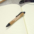 A black plastic pen with a cork grip rests on an open notebook with lined pages. It features a logo.
