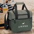 A green cooler bag with a textured front, featuring a shoulder strap and handles, positioned near a drink bottle on sand.