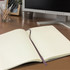 A large, soft cover Moleskine notebook in cream with lined pages and a purple ribbon bookmark, placed on a desk.