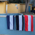 Three cotton beach towels hang over a stone ledge by a pool, featuring navy, black, red, and white stripes.
