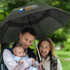 A black umbrella with a logo, held by a man carrying a baby, while a woman smiles in a park setting.