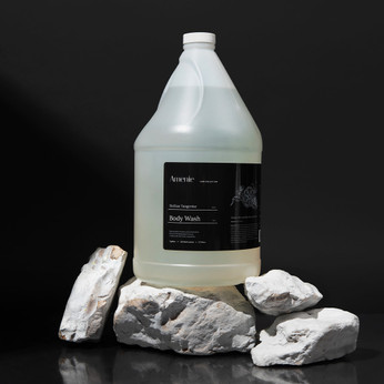 Amenie Sicilian Tangerine Body Wash styled with bath towel and white spa stones on a dark green fabric