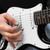 Close-up of an electric guitar being played with a blue pick, showing strings, pickups and white pickguard.