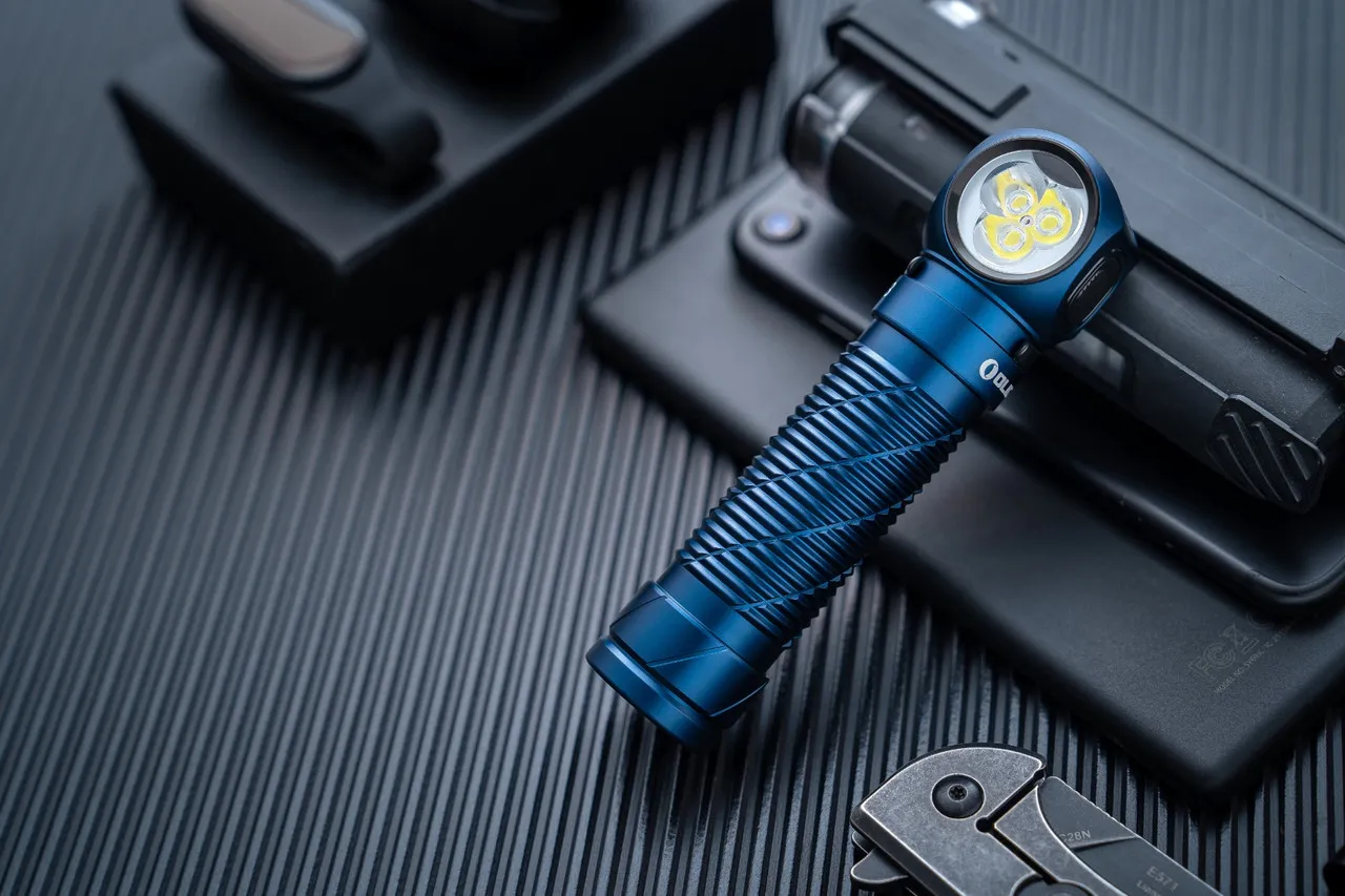Blue Olight Perun 3 High Performance 3000 lumen rechargeable headlamp lying on a dark surface with other tech accessories.