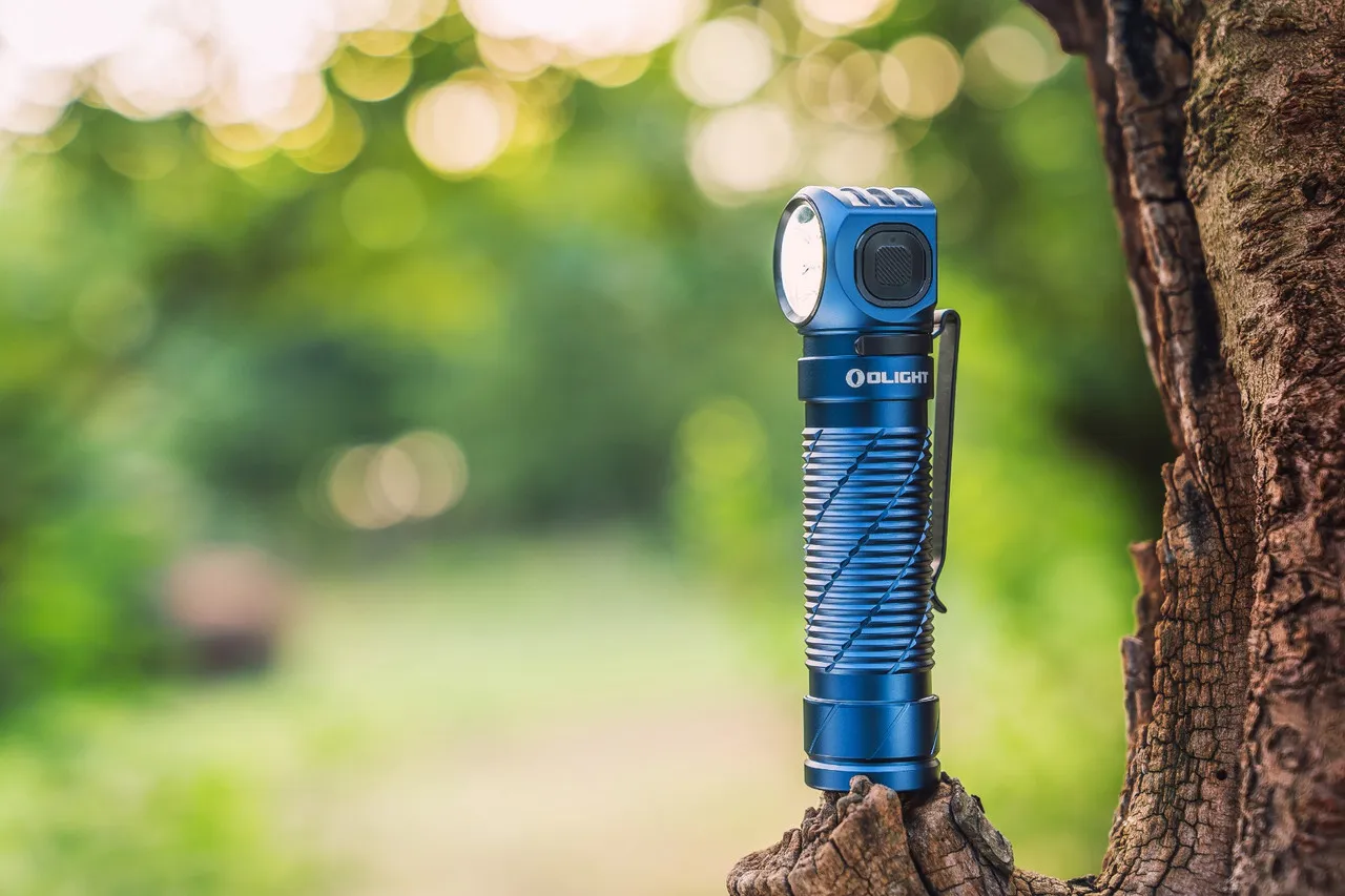 Blue Olight Perun 3 High Performance 3000 lumen rechargeable headlamp standing upright on a tree stump outdoors.