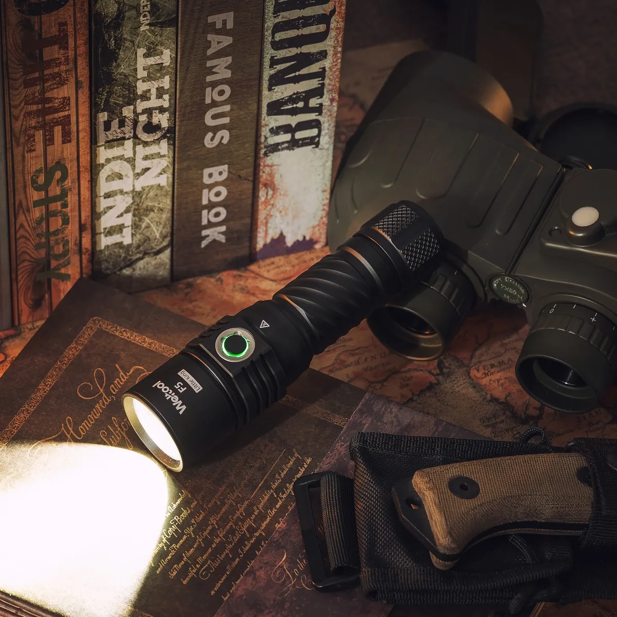 Weltool F5 flashlight illuminating a desk setup with vintage books, binoculars, and a tactical knife.
