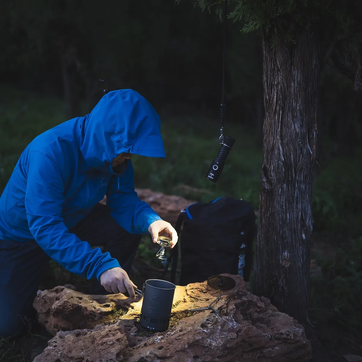 Weltool F5 flashlight hanging from a tree branch, illuminating a person cooking outdoors at a campsite at night.