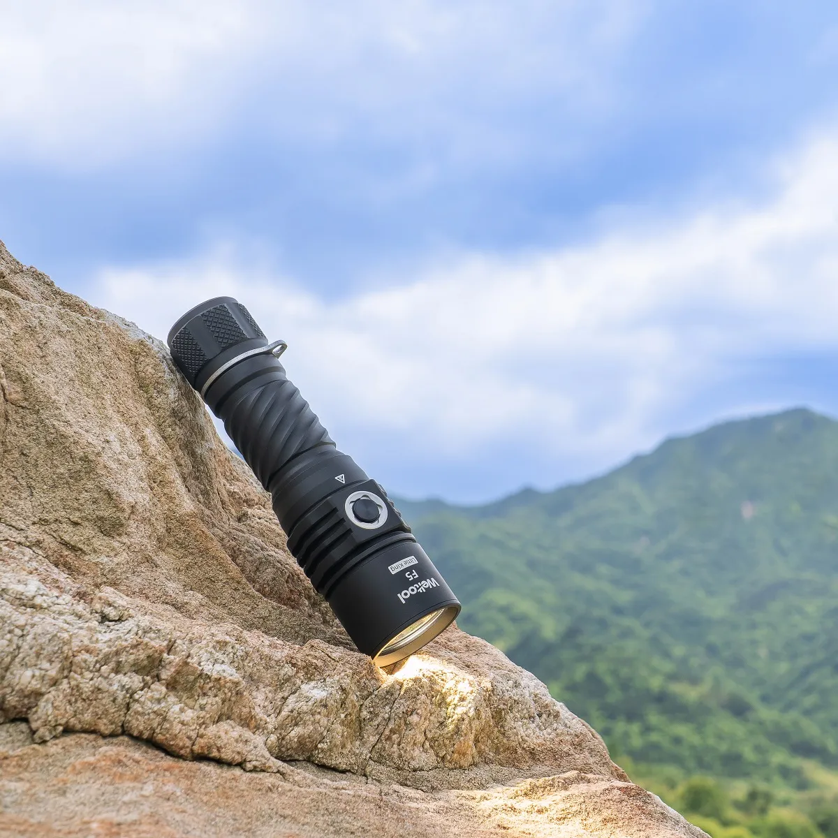 Weltool F5 flashlight resting on a textured rock outdoors with mountains and sky in the background.