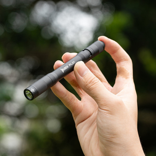 Wuben PL01 penlight flashlight held in a hand outdoors against a blurred green background.