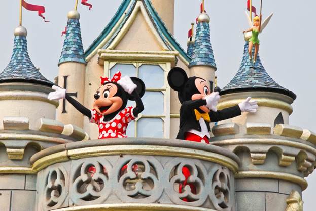 Mickey Mouse and Minnie Mouse wave from a castle balcony at a Disney park.