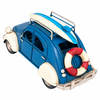 Rear view of a vintage blue Volkswagen Beetle-inspired metal car featuring a red and white life preserver, blue and white surfboard, metal roof rack, cream fenders, and distressed coastal-inspired finish.
